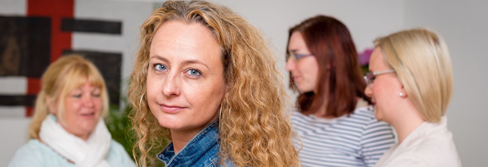 Une femme aux cheveux bouclés regarde la caméra, trois autres femmes discutent en arrière-plan.
